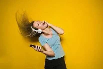 A woman in headphones dancing and singing with her phone against a yellow wall