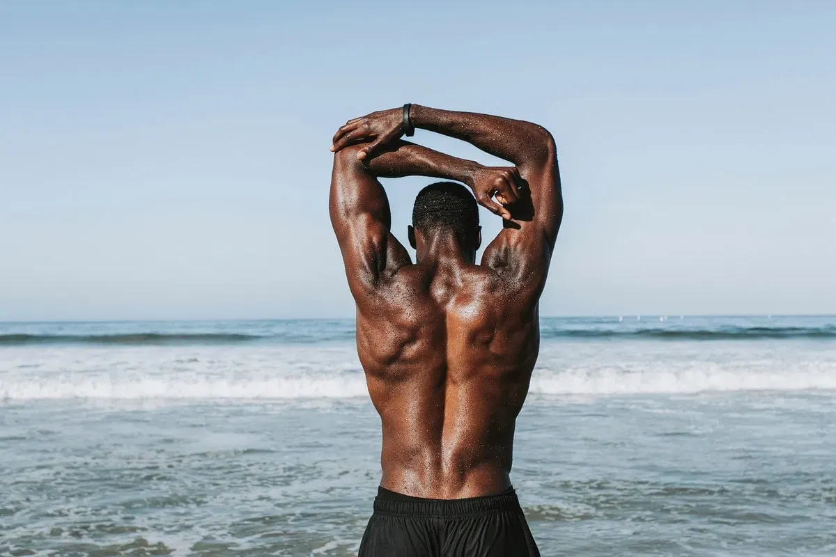 Homme torse nu de dos, les bras levés derrière la tête, face à la mer