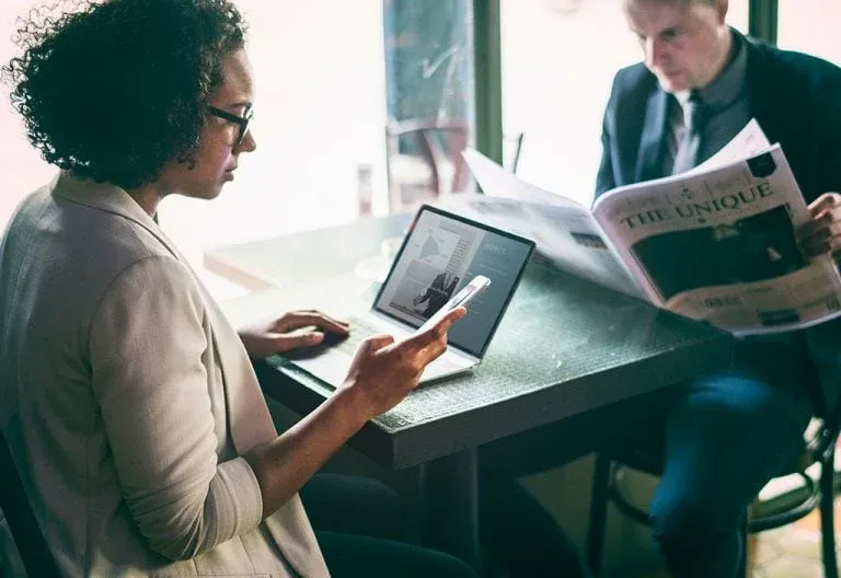 A woman checks her phone at a laptop in a cafe as a man beside her reads a paper